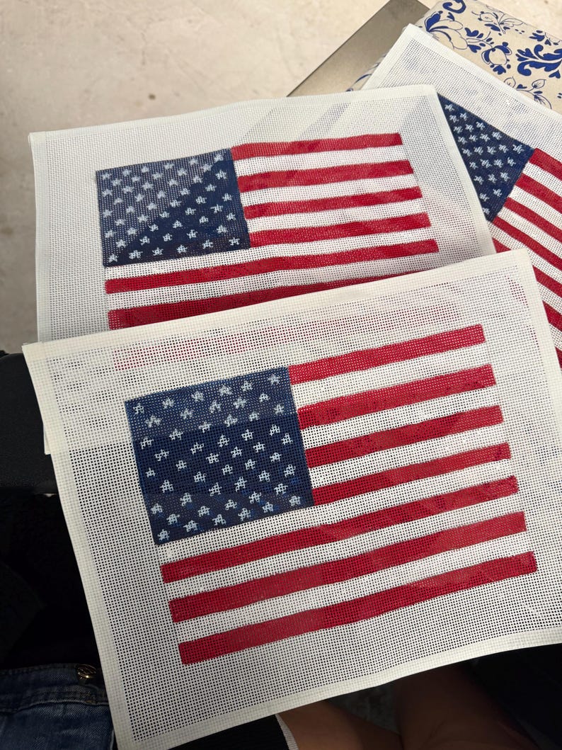 May include: Two rectangular needlepoint canvases featuring a printed American flag design. Each canvas displays a blue field with white stars and alternating red and white horizontal stripes on a white mesh grid background, ready for embroidery.