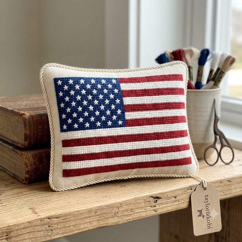 May include: A rectangular needlepoint pillow featuring the American flag design with a blue canton of white stars and alternating red and white stripes. The pillow is edged with a thick cream-colored twisted rope trim and has a tan tag attached.