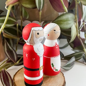 May include: A pair of hand-painted wooden Christmas decorations featuring Santa Claus and Mrs Claus. Santa is wearing a red suit with white trim and a white beard. Mrs Claus is wearing a red dress with a white apron and a green holly sprig.
