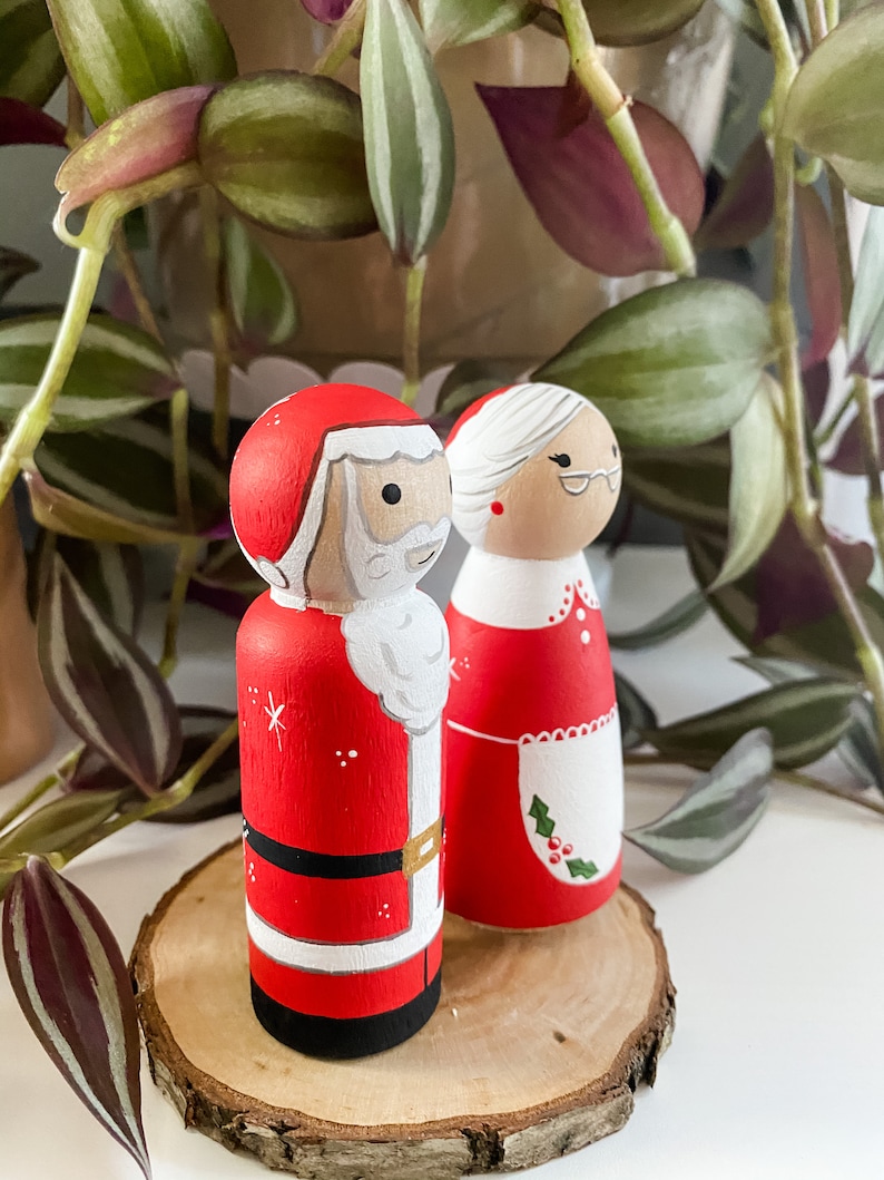 May include: A pair of hand-painted wooden Christmas decorations featuring Santa Claus and Mrs Claus. Santa is wearing a red suit with white trim and a white beard. Mrs Claus is wearing a red dress with a white apron and a green holly sprig.