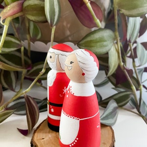 May include: A pair of hand-painted wooden Christmas decorations featuring Santa Claus and Mrs Claus. Santa is wearing a red suit with white trim and a black belt. Mrs Claus is wearing a red dress with a white apron and white trim. The decorations are standing on a wooden slice.