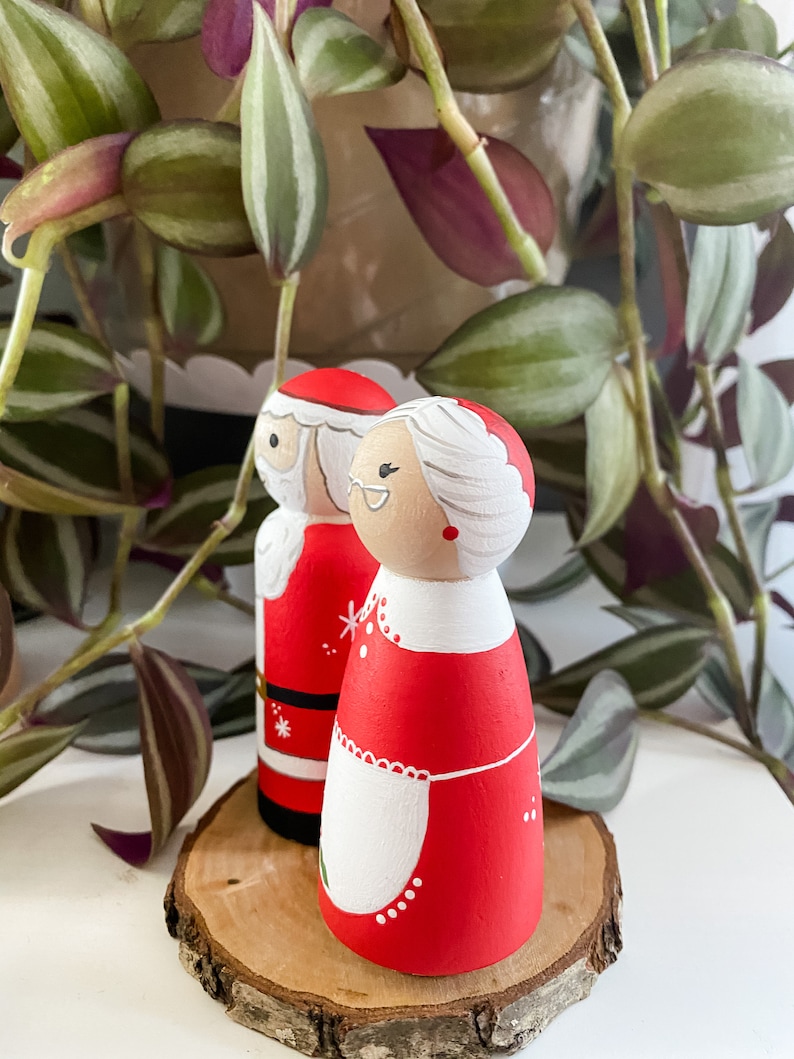 May include: A pair of hand-painted wooden Christmas decorations featuring Santa Claus and Mrs Claus. Santa is wearing a red suit with white trim and a black belt. Mrs Claus is wearing a red dress with a white apron and white trim. The decorations are standing on a wooden slice.
