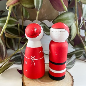 May include: A pair of hand-painted wooden peg dolls dressed as Santa and Mrs Claus. The dolls are standing on a wooden slice. The Santa doll is wearing a red suit with a white trim and a black belt. The Mrs Claus doll is wearing a red dress with a white bow and a white hat.