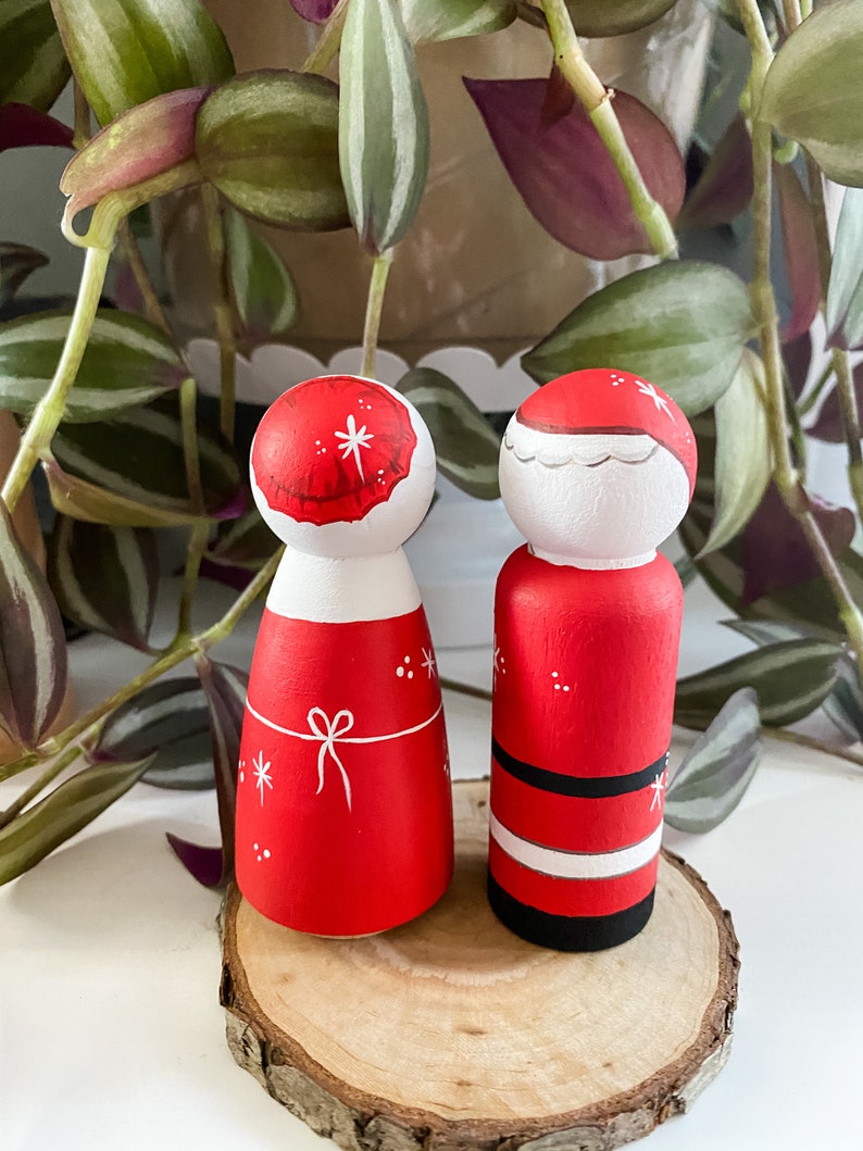 May include: A pair of hand-painted wooden peg dolls dressed as Santa and Mrs Claus. The dolls are standing on a wooden slice. The Santa doll is wearing a red suit with a white trim and a black belt. The Mrs Claus doll is wearing a red dress with a white bow and a white hat.