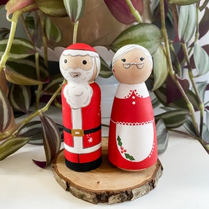 May include: A pair of hand-painted wooden Christmas decorations featuring Santa Claus and Mrs Claus. Santa is wearing a red suit with white trim and a white beard. Mrs Claus is wearing a red dress with a white apron and a white hair bun.