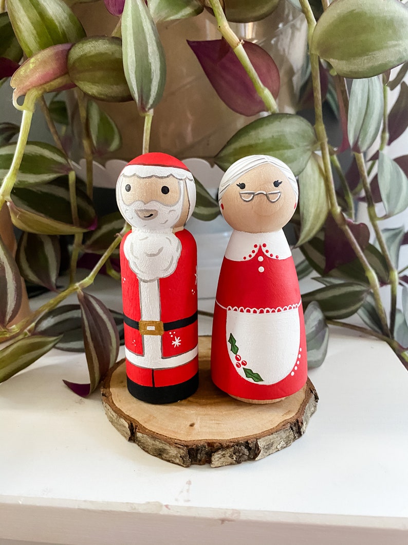 May include: A pair of hand-painted wooden Christmas decorations featuring Santa Claus and Mrs Claus. Santa is wearing a red suit with white trim and a white beard. Mrs Claus is wearing a red dress with a white apron and a white hair bun.