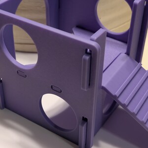 May include: A light purple modular pet hideout constructed from flat panels with circular cutouts on each side. The structure features an integrated ramp with horizontal ridges leading to an elevated platform, all assembled with interlocking joints.