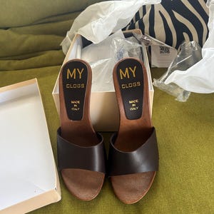 May include: A pair of dark brown leather clogs with wooden soles and open toes. The footbed features a black label with gold text that reads MY CLOGS MADE IN ITALY. The clogs are presented in a white cardboard box with white tissue paper.