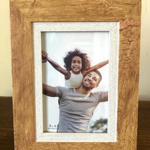 Puede incluir: Un marco de fotos rectangular con un acabado de grano de madera marrón claro natural y un borde interior blanco. El marco contiene una fotografía de un adulto sonriente cargando a un niño sobre sus hombros. El texto 10,1 cm x 15,2 cm está impreso.