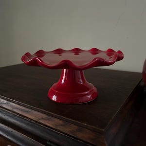 May include: A glossy red ceramic cake stand featuring a circular top with a decorative scalloped, wavy edge. The stand is supported by a sturdy, flared pedestal base, providing a raised platform for displaying baked goods or other items.