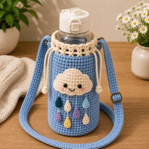 May include: A clear plastic water bottle with a white locking lid, encased in a light blue crocheted sleeve. The sleeve features a cream-coloured crocheted cloud with a smiling face and dangling multi-coloured raindrops, plus a matching strap.