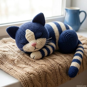 以下が含まれることがあります： 眠っている表情のクロシェ編みの猫のぬいぐるみ。閉じた目、ピンクの鼻、長い白いひげが特徴です。体、頭、尻尾にはダークブルーとクリーム色のストライプ模様があり、顔と胸元はクリーム色で仕上げられています。