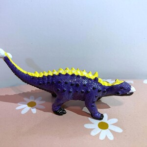 May include: A small dinosaur figurine featuring a textured purple body with black spots and a bright yellow ridge running along its back. The creature has a long tail ending in a white bulbous tip and a white snout, standing in a quadrupedal pose.