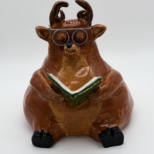 May include: A glossy brown ceramic figurine of a seated deer with antlers, wearing wire-rimmed glasses and holding an open green book. The deer has black hooves and a smooth, reflective finish, depicted in a reading pose against a plain background.