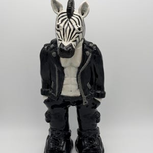 May include: A ceramic figurine of a zebra with a black and white striped head, wearing a black leather-style jacket over a bare chest, black trousers, and black shoes. The figure stands upright with hands in pockets, featuring a glossy finish.