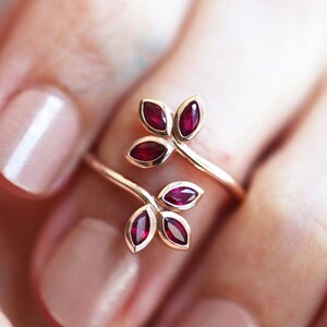 May include: A bypass ring crafted from a polished gold-tone metal band, featuring two leaf-shaped motifs at each end. Each leaf cluster contains three marquise-cut deep red gemstones, arranged to resemble a small branch or floral sprig design.