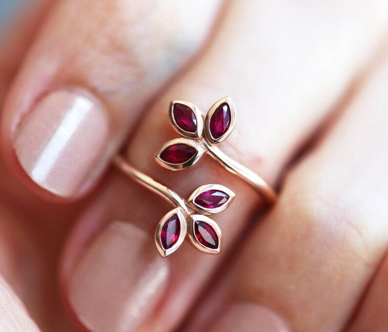 May include: A bypass ring crafted from a polished gold-tone metal band, featuring two leaf-shaped motifs at each end. Each leaf cluster contains three marquise-cut deep red gemstones, arranged to resemble a small branch or floral sprig design.