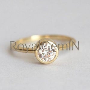 May include: A gold ring featuring a single round, clear, faceted gemstone set in a smooth, circular bezel setting. The band is thin and polished gold. The text RoyalGemIN is superimposed across the centre of the ring in a dark grey font.