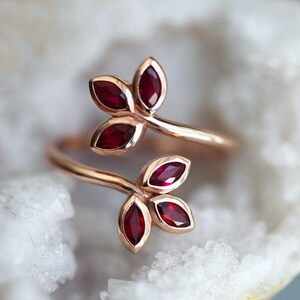 May include: A bypass ring crafted from rose-toned metal, featuring two leaf-shaped motifs at each end. Each leaf motif contains three marquise-cut deep red gemstones arranged in a cluster, creating a botanical design that wraps around the finger.