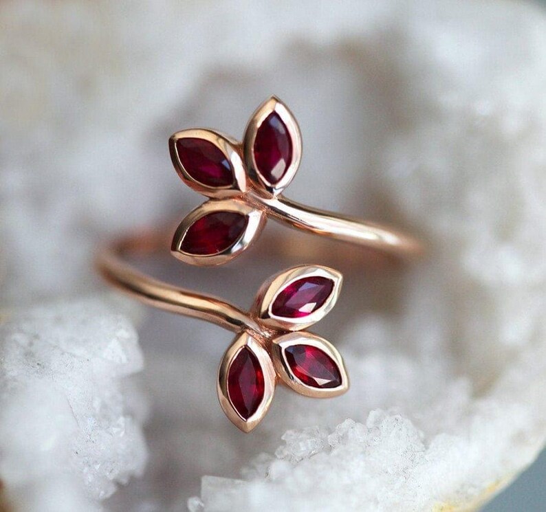 May include: A bypass ring crafted from rose-toned metal, featuring two leaf-shaped motifs at each end. Each leaf motif contains three marquise-cut deep red gemstones arranged in a cluster, creating a botanical design that wraps around the finger.