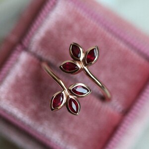 May include: A bypass ring crafted from rose-toned metal, featuring two leaf-shaped motifs at each end. Each leaf cluster contains three marquise-cut deep red gemstones set in smooth bezels, creating a botanical design that wraps around the finger.