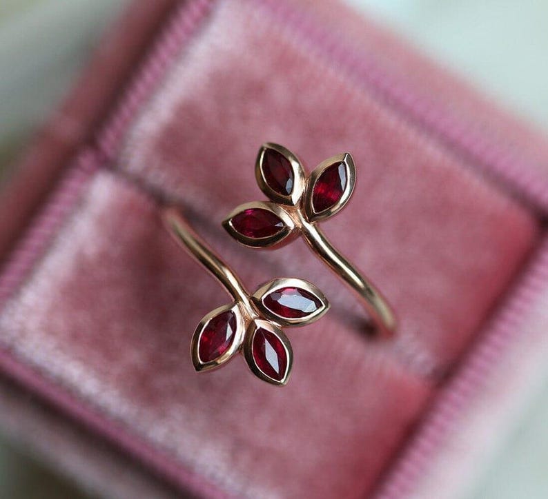 May include: A bypass ring crafted from rose-toned metal, featuring two leaf-shaped motifs at each end. Each leaf cluster contains three marquise-cut deep red gemstones set in smooth bezels, creating a botanical design that wraps around the finger.