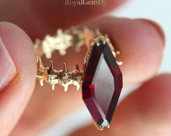 Könnte beinhalten: Ein Goldring mit einem großen, länglichen, drachenförmigen, tiefroten Edelstein. Die Ringschiene hat ein strukturiertes, wirbelsäulenartiges Design mit hervorstehenden Spitzen. Der Text RoyalGemIN ist oben im Bild zu sehen.