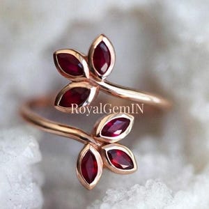 May include: A bypass ring crafted from rose-coloured metal featuring four marquise-cut deep red rubies arranged in two leaf-like clusters. The text RoyalGemIN is centred between the two ends of the ring band, which curves to form the floral design.