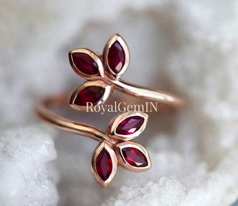 May include: A bypass ring crafted from rose-coloured metal featuring four marquise-cut deep red rubies arranged in two leaf-like clusters. The text RoyalGemIN is centred between the two ends of the ring band, which curves to form the floral design.