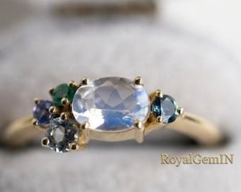 Könnte beinhalten: Ein Goldring mit einem großen, oval geschliffenen Mondstein als Mittelstein, akzentuiert durch eine Ansammlung kleinerer, rund geschliffener Edelsteine in Blau-, Grün- und Grautönen. Der Text RoyalGemIN ist unten rechts im Bild zu sehen.