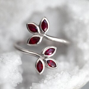 May include: A silver bypass ring featuring two leaf-shaped motifs, each set with three marquise-cut deep red rubies. The metal band is polished and smooth, with the two ends of the ring curving outwards to display the ruby-encrusted leaf designs.