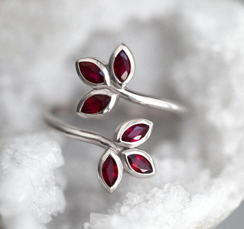 May include: A silver bypass ring featuring two leaf-shaped motifs, each set with three marquise-cut deep red rubies. The metal band is polished and smooth, with the two ends of the ring curving outwards to display the ruby-encrusted leaf designs.