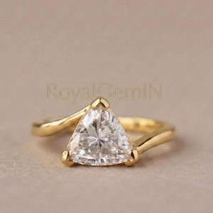 May include: A gold ring featuring a trillion-cut clear gemstone set in a bypass-style band. The metal has a polished finish, and the stone is faceted to reflect light. The text RoyalGemIN is visible in the background behind the ring.