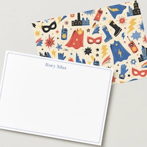 May include: A rectangular white stationery card with a thin blue border and the name Rory Silas printed in blue text. Behind it is a patterned envelope liner featuring colorful superhero-themed illustrations including capes, masks, and cityscapes.