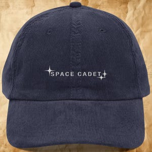 May include: A navy blue corduroy baseball cap featuring white embroidered text that reads SPACE CADET, flanked by two four-pointed star icons on each side. The cap has a structured crown, a curved brim, and a button at the top center.