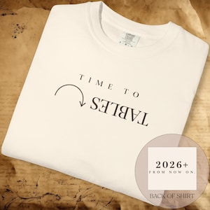 May include: A cream-colored t-shirt featuring the black text TIME TO and the word TABLES printed upside down with a curved arrow pointing toward it. A circular graphic overlay displays the text 2026+ FROM NOW ON. BACK OF SHIRT.