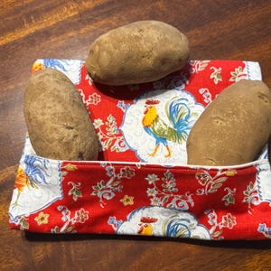 May include: Three brown potatoes rest in a red and white fabric pouch. The pouch features a rooster and floral pattern. The potatoes are of varying sizes and are placed in the pouch to be stored.