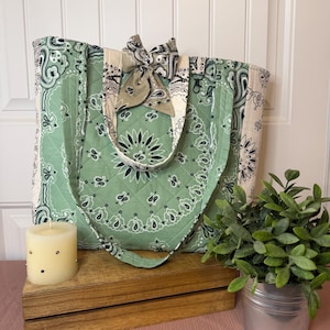 May include: A quilted tote bag featuring a sage green and cream bandana print with a black paisley pattern. The bag includes two matching fabric handles and a decorative bow attached to the top center, with a small black metal clasp visible.