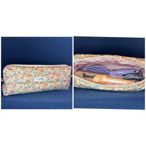 May include: A quilted rectangular zippered pouch featuring a yellow floral pattern with pink, blue, and green flowers. A white label reads Handmade by Piece Together. The interior contains a light purple hairbrush and a peach-colored hair tool.
