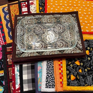 May include: A collection of colorful quilted pot holders and trivets in various patterns. A clear glass baking dish sits on a paisley-patterned trivet. Other designs include chickens, fruit, and floral motifs. The items are arranged on a dark surface.