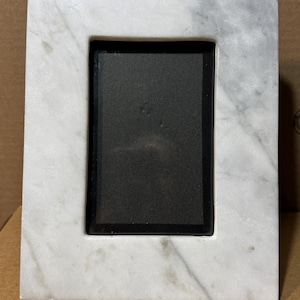 May include: A rectangular picture frame featuring a white marble border with natural grey veining. The frame has a smooth, polished finish and a centered, dark black rectangular insert, providing a stark contrast to the light stone material.