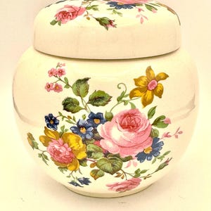 May include: A cream-colored ceramic jar with a matching lid, featuring a colorful floral pattern of pink roses, yellow daffodils, and small blue flowers. The rounded vessel has a smooth, glossy finish and a wide body with a tapered base.