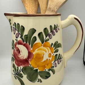 May include: A cream-colored ceramic pitcher featuring hand-painted floral motifs in shades of red, yellow, and green, with small purple accents. The pitcher has a curved handle and a brown rim, and it contains three light-colored wooden kitchen utensils.