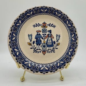 May include: A round ceramic plate with a cream-colored base featuring a central illustration of two figures in traditional attire standing beside a floral motif. The rim is decorated with a dark blue repeating pattern of stylized leaves and flowers.