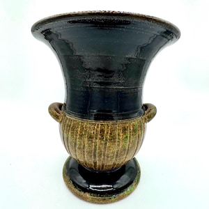 May include: A ceramic urn-shaped vase featuring a glossy black upper section and a textured, ribbed lower body in mottled olive green and brown tones. The vessel includes two small side handles and a wide, flared rim, set on a circular base.