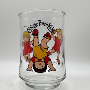May include: A clear glass tumbler featuring a colorful graphic of three Cabbage Patch Kids characters. One character is upside down in the center wearing yellow overalls and a red shirt, flanked by two characters in red outfits. The text Cabbage Patch Kids is printed in black above the figures.