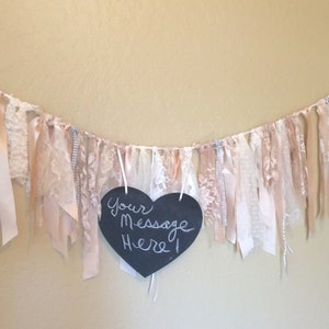 May include: A white and blush pink fabric garland with a black heart-shaped chalkboard sign that says "Your Message Here!" hanging from it.