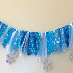 May include: A blue and white fabric banner with snowflake cutouts. The banner is made of various shades of blue and white tulle and ribbon. It is strung on a white cord and has silver glitter snowflakes.