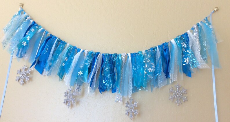 May include: A blue and white fabric banner with snowflake cutouts. The banner is made of various shades of blue and white tulle and ribbon. It is strung on a white cord and has silver glitter snowflakes.