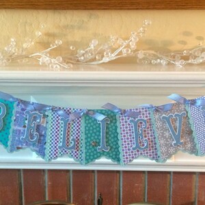 May include: A blue and white banner with the word "BELIEVE" spelled out in silver letters. The banner is made of paper and has a scalloped edge. It is hanging from a white string.
